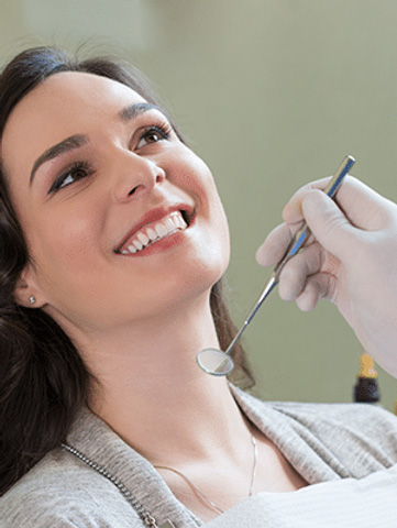 The image shows a woman sitting in a dental chair with her eyes closed, smiling broadly, while receiving dental care from a professional who is working on her teeth.