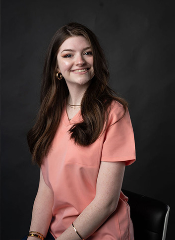 The alt text for this image could be A young woman with long brown hair wearing a pink blouse, sitting in front of a black background.