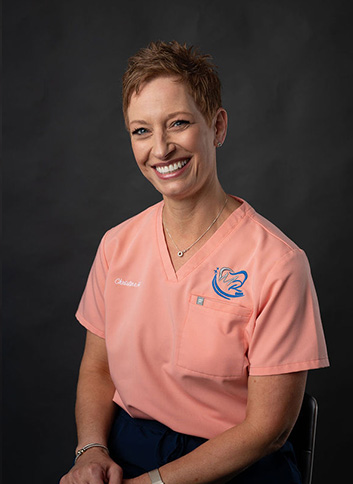 A woman wearing a pink scrub top with a logo on the left chest area, smiling at the camera.