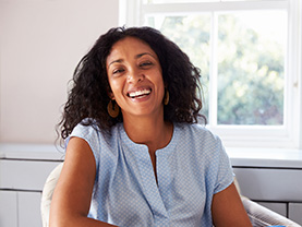 The image shows a woman smiling at the camera, wearing a light blue top and seated on a chair with a window behind her.