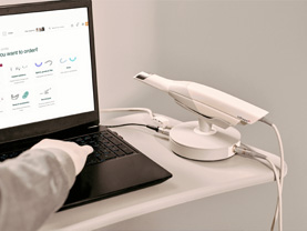 The image depicts a person interacting with a laptop screen displaying a webpage, with a focus on dental care products such as toothbrushes and an electric toothbrush holder, which are also physically present in the scene.