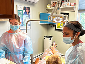 Two dental hygienists wearing face masks and protective gowns, working at a dental station with a patient seated in a chair.