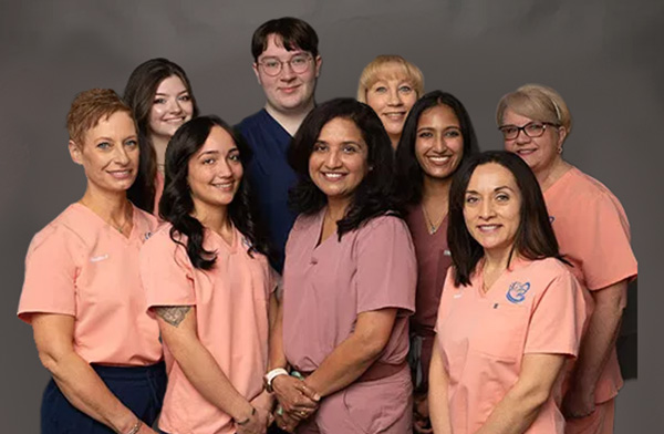 The image shows a group of people, likely nurses, standing together with a young man in front, all wearing scrubs with a color scheme that includes shades of pink and purple.